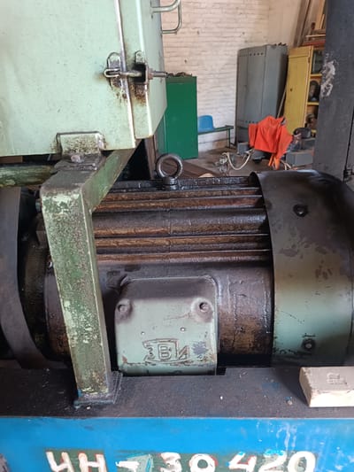 The image shows a close-up of industrial equipment, likely a motor, in what appears to be a workshop or storage area. The motor casing is dark and shows signs of wear and oil. A light green metal housing is attached to the top and left of the motor, also appearing weathered. In the background, there are various metal cabinets, shelves, and what looks like a piece of orange fabric. The environment suggests an industrial or manufacturing setting. The floor is dark and appears to be concrete or a similar material. The image is well-lit, highlighting the textures and details of the machinery. There is no indication of the specific location within Vuhlehirsk, Ukraine, beyond it being an industrial setting.