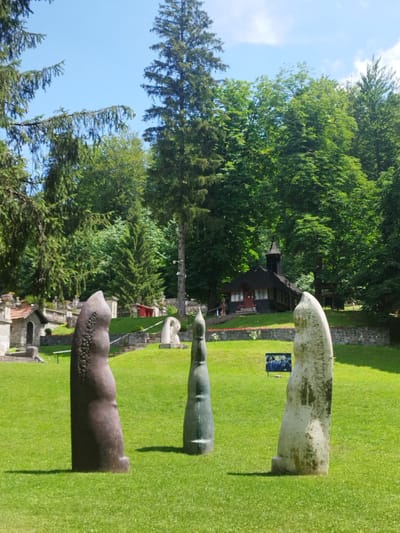 The image depicts an outdoor art installation in the city of Bușteni, Romania, specifically within the Cantacuzino Castle gardens. In the foreground, three abstract stone sculptures stand on a lush green lawn. The sculpture on the left is dark brown with a textured surface, appearing to be made of bronze or a similar dark metal. The central sculpture is slender and dark green, with a smooth, tapered form. The sculpture on the right is lighter in color, appearing weathered and perhaps made of a lighter stone or concrete. In the middle ground, a small, dark wooden building resembling a chapel or pavilion with a steeple is visible. Stone staircases and retaining walls lead up a gentle slope behind the chapel, surrounded by dense green trees. A framed picture displaying three individuals in formal attire is placed on the lawn in front of the chapel. The sky is clear blue with a few scattered white clouds, and sunlight casts shadows on the grass. The overall impression is a serene and artistic park setting.