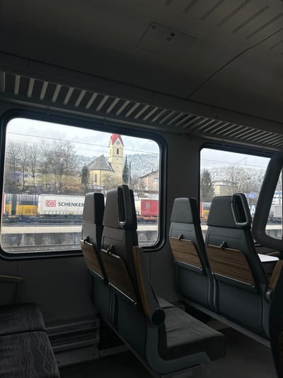 The image depicts the interior of a passenger train car, looking outward through a window at a railway environment adjacent to the city of Kufstein, Austria.
Inside the train, multiple rows of unoccupied grey passenger seats with wood-grain accent panels are visible. The ceiling features light-colored panels with linear ventilation grilles and overhead fixtures. A heater unit is positioned on the floor beneath the window.
Outside the window, multiple parallel railway tracks are present. Several freight cars are parked on these tracks, including a white "DB SCHENKER" branded car, a yellow car, and a red car further down the line. Beyond the railway, the prominent Stadtpfarrkirche St. Vitus, identifiable by its yellow facade and distinctive red-capped steeple, stands on an elevated section of the city. Additional buildings, including multi-story structures, are visible around the church. In the background, snow-dusted mountains or hills rise under an overcast sky, with bare deciduous trees and some evergreens dotting the landscape. No individuals are visible inside or outside the train.