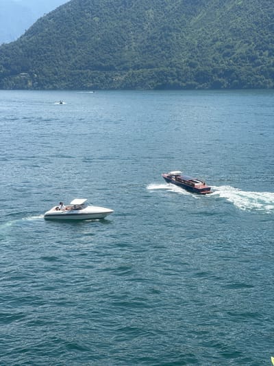 This is a high-angle, full shot of a lake in Tremezzina, Italy, with a heavily wooded mountain range in the background. Two boats are visible on the water. In the foreground left, a white motorboat with a canopy top is moving to the left, leaving a wake. Three individuals are seated in the boat. In the middle ground right, a dark blue motorboat with a red stripe and a white canopy is moving to the right, also leaving a wake. The water is choppy with light reflecting off the surface. The mountain range is covered in dense green trees and appears steep. The sky is clear and bright.