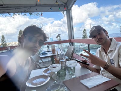 The image captures two males seated at an outdoor restaurant table in Bondi Beach, Australia, under a partly cloudy sky. The male on the left, in the foreground, has dark hair, a mustache, wears dark sunglasses and a dark sleeveless top; a significant lens flare obscures the lower half of his face and upper chest. The male on the right has dark hair, a mustache, wears a dark baseball cap with a white 'N' logo, a light-colored short-sleeved collared shirt with an emblem on the left chest, and a watch or bracelet on his left wrist. He holds a dark smartphone in his left hand and displays a 'peace' sign with his right hand, looking towards the camera.

The white table between them holds two empty oval plates, two clear glass goblets with salted rims containing residual liquid, a clear glass bottle, a small paper cup, chopsticks in a holder, and a brown menu folder with a white napkin. Overhead, string lights with multicolored bulbs are visible against the sky. The restaurant features a glass-paneled railing with reddish-brown framing, providing a view of the external environment. Beyond the railing, a street with a white parked SUV, evergreen trees (likely Norfolk Pines), and the ocean with breaking waves are visible under a bright, cloud-dotted sky, characteristic of a coastal urban area in Bondi Beach.