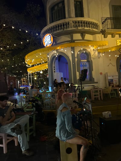 The image shows a nighttime scene at a restaurant or bar in Mazatlán, Mexico. A yellow awning with lights covers an outdoor seating area. People are seated at tables, and others are standing and interacting. The restaurant is decorated with string lights in the trees. The building has architectural details, including arched windows and a rounded tower. A sign with the restaurant's name is visible. People are casually dressed. Some patrons are seated in chairs. It is a lively social atmosphere.