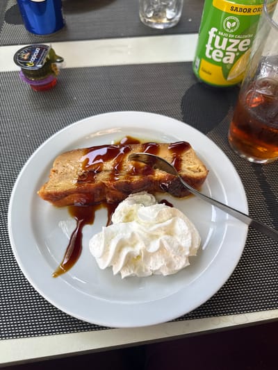 A close-up, eye-level shot captures a dessert on a white plate, positioned on a dark grey placemat. The dessert is a slice of bread pudding, generously drizzled with a dark caramel sauce. To the right of the pudding, a dollop of whipped cream sits on the plate. A silver spoon is embedded in the bread pudding, indicating it is being eaten. In the background, out of focus, a clear glass filled with a dark amber liquid, likely iced tea, and a green can of "Fuze Tea" are visible. Also present is a small plastic condiment cup with a purple lid, and a portion of a blue can. The overall scene suggests a casual dining setting, possibly a cafe or restaurant in Getafe, Spain, where dessert and beverages are being consumed.