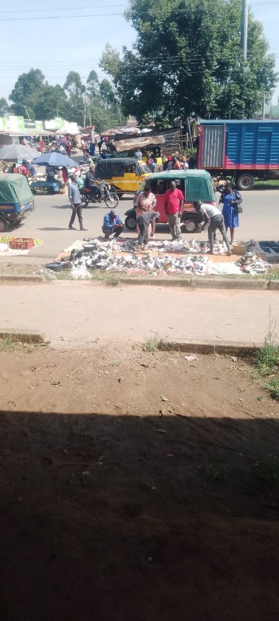 This image depicts an active roadside market scene in Isukha North ward, Kenya, observed on a clear, sunny day. The foreground is partially obscured by a dark shadow, revealing a dirt verge with sparse vegetation and a concrete curb.
On the paved roadside, a significant display of footwear, consisting of numerous shoes, sandals, and slippers, is laid out on multiple light-colored tarpaulins. Approximately five individuals are actively engaged around this display; one person crouches examining items, while others stand, some appearing to interact with the merchandise. Small dark containers are visible near the footwear. To the left of this display, a red plastic crate holds green and red produce.
On the road, vehicle and pedestrian traffic is evident. A green three-wheeled auto-rickshaw (tuk-tuk) is parked near the footwear display, with at least two occupants visible and one person standing next to it. A yellow tuk-tuk is further down the road, and a motorcyclist is also present. Pedestrians are walking along the road; notable individuals include a man in a blue shirt walking left and a woman in a blue dress standing near the green tuk-tuk. A blue market umbrella shades goods further left on the roadside.
The background reveals a bustling market environment with numerous temporary stalls and permanent structures featuring corrugated iron and tarpaulin roofs. Many people are visible within this market area. A large blue truck with pink detailing is parked on the right side of the road. Overhead, power lines crisscross the frame. Tall, leafy trees dominate the distant background against a clear blue sky, with some buildings also discernible.
