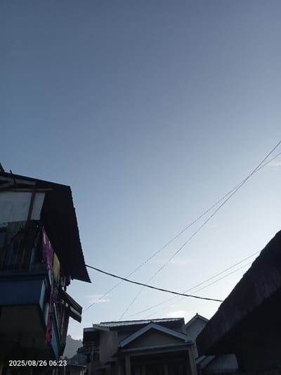 The image presents an upward view of a residential neighborhood in Cibeureum, Indonesia, captured on 2025/08/26 at 06:23. The upper half of the frame is filled by a clear, light blue sky with a subtle gradient from darker blue at the top to lighter blue near the horizon, where faint, wispy clouds are visible. In the lower-left, a section of a multi-story building is prominent, featuring a dark, corrugated metal roof and a white wall. Below the roofline, a balcony or exterior drying area displays several items of hanging laundry, including purple and yellow textiles. In the mid-ground, a cluster of residential buildings with pitched roofs is present. One structure in the lower-center exhibits a light-colored facade and a distinctive gabled roof section above an entrance. Multiple thin, dark utility wires crisscross the sky, with one thicker, dark cable running horizontally across the lower-mid portion of the image. The distant background reveals faint outlines of hills or mountains shrouded in haze. No individuals are visible.