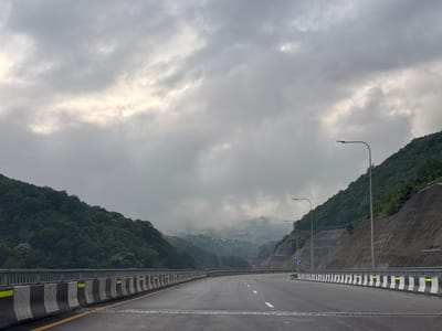 The image shows a view of a highway curving through a mountainous, forested landscape under a cloudy sky. The highway has a guardrail made of concrete barriers with alternating black and white stripes and yellow reflective markers. There are streetlights lining the highway. The surrounding mountains are covered in dense green trees, and the further peaks are shrouded in mist, with some buildings and structures visible on a distant hilltop. The road surface appears wet, suggesting recent rain. The scene is devoid of people or vehicles, creating a sense of quietness. This highway is located near Tsakva, Georgia.