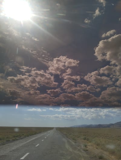 The image depicts a road stretching through a vast, flat, grassy plain under a dramatic sky. The road, paved with asphalt and marked with white dashed lines, appears to lead into the distance. The landscape is characterized by dry, yellowish-brown grass. In the far distance, mountains are visible on the horizon. The sky is a mix of dark gray clouds and patches of bright blue sky, with a prominent sun creating lens flares and rays of light. There are no people or vehicles clearly visible on the road or in the immediate vicinity. The scene evokes a sense of open space and remoteness.