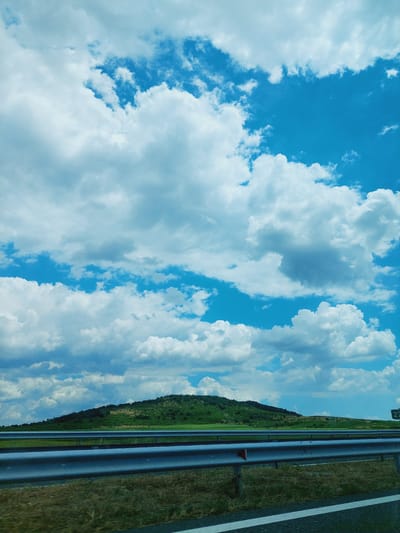 The image shows a view from a road in the vicinity of Drazhevo, Bulgaria, under a bright blue sky filled with white, fluffy clouds. In the foreground, a highway guardrail runs horizontally across the frame, with dry grass and a portion of the road visible below. Beyond the guardrail, a rolling green hill is covered with trees and vegetation. The sky dominates the upper portion of the image, with a dramatic display of cumulus clouds. A highway sign with the number "27" is visible on the right side of the image.