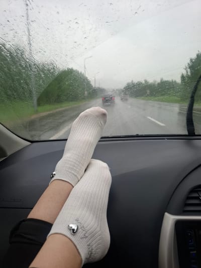 Two feet, adorned with white ribbed socks that each feature a silver heart charm, are resting on the dashboard of a car. The perspective suggests the photo was taken from the passenger seat. Outside the car, a rainy day is depicted. The windshield is streaked with rain, obscuring the view of a road with cars driving in the distance. Green trees line the side of the road, and the sky is overcast and gray. The interior of the car is visible in the foreground, with the dark dashboard dominating the lower portion of the frame. The location specified, Malaya Elanka, Russia, is a rural area, and the scene is consistent with driving on a wet road in such a setting.