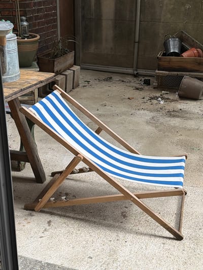 A deckchair with blue and white stripes stands on a textured grey surface. A wooden planter box and various containers are nearby. The scene appears to be outdoors, possibly in a courtyard or backyard. The location is unknown, and it is not possible to determine the city of Aubervilliers, France from this image alone. There are no people in the frame.