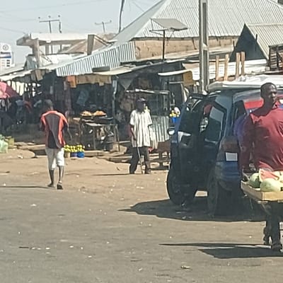 The image depicts a daytime street scene in a market area of Haifam, Nigeria. In the immediate foreground on the right, a dark-skinned male wearing a red short-sleeved shirt is partially visible, carrying a wooden tray with multiple green watermelons on his left shoulder. He faces left, appearing to walk forward. To his left, a dark-colored, compact vehicle, possibly a three-wheeled passenger vehicle or a small car, is parked with its driver's side door ajar. Behind this vehicle, slightly to the left, a dark-skinned male stands facing right, wearing a white and dark-striped short-sleeved shirt and dark trousers, holding a slender stick or cane in his right hand. Further left in the midground, another dark-skinned male, dressed in a reddish-orange shirt with a tan back panel and light shorts, walks away from the viewer towards the left. The ground in the mid-distance is unpaved, dusty, and uneven, while the immediate foreground shows a paved road surface. In the background, a row of market stalls or shops with corrugated metal roofs and various building materials like wood and fabric are visible. Several stalls display goods, including what appear to be fried items on trays and yellow fruits (possibly plantains or bananas) in blue containers. Overhead power lines and utility poles are present in the upper background against a clear, bright sky, indicating strong sunlight casting shadows on the ground.