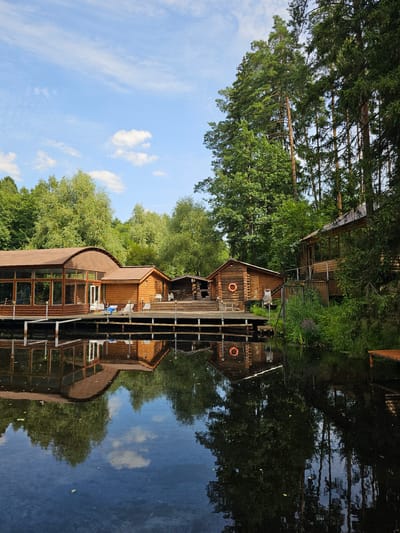 In the city of Ulitkino, Russia, a tranquil waterside scene unfolds. Several wooden buildings, featuring rustic log construction and brown roofs, are situated on a wooden deck extending over a dark body of water. The largest building on the left has a distinctive curved roof and large glass windows. Adjacent to it are smaller log cabins, one of which displays an orange life preserver on its exterior. The buildings are surrounded by lush green trees, with tall pine trees dominating the background on the right. The water perfectly reflects the buildings and the surrounding foliage, creating a mirror image that adds to the serene atmosphere. The sky above is a clear blue, dotted with a few white clouds. The scene appears to be a resort or recreational area, possibly a spa or a lodge, given the presence of lounge chairs and the overall ambiance. There are no people visible in the image.