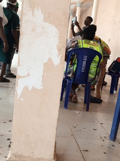 The image captures an indoor or covered outdoor area in Ajalli, Nigeria. In the immediate foreground, a cream-colored vertical structural pillar exhibits extensive peeling paint, revealing lighter patches underneath. To the left of this pillar, multiple individuals are partially visible standing, including one person in a dark green uniform-like garment. To the right of the foreground pillar, a person is seated on a blue plastic chair, facing away from the viewer. This individual wears a neon yellow-green reflective safety vest, and the chair's back displays faded white text, possibly "RENTALS". Their feet, clad in light-colored flip-flops, are visible beneath the chair. Behind the seated person, another individual, also wearing a reflective vest, is partially visible. Further to the right and in the background, a person with short, dark hair stands, holding a black smartphone horizontally, seemingly operating it for recording or photography. The floor consists of light-colored square tiles, which are marked by scattered dark debris and small paper fragments. In the background, decorative white columns with capitals are visible, suggesting the architecture of a public or semi-public building. A dark bag or object rests on the tiled floor near the chairs.