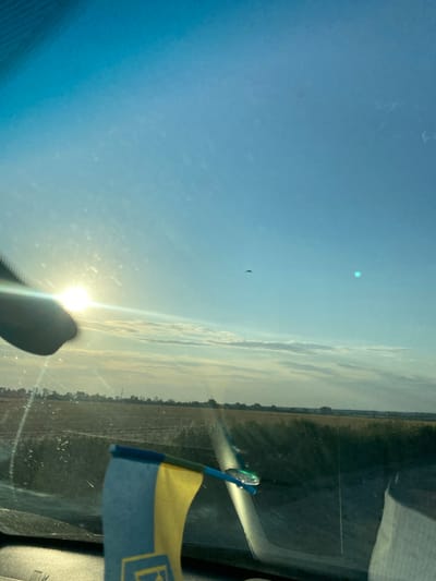 A Ukrainian flag is visible in the foreground of the image, attached to the interior of a vehicle. The flag is positioned to the left side of the frame. The view from the vehicle shows a sunlit landscape with a field under a bright blue sky. There are a few clouds scattered across the sky, and a distant bird can be seen flying. The sun is a bright, prominent light source, casting a glare on the vehicle's windshield. The overall scene suggests an outdoor environment, likely rural, in Ukraine.
