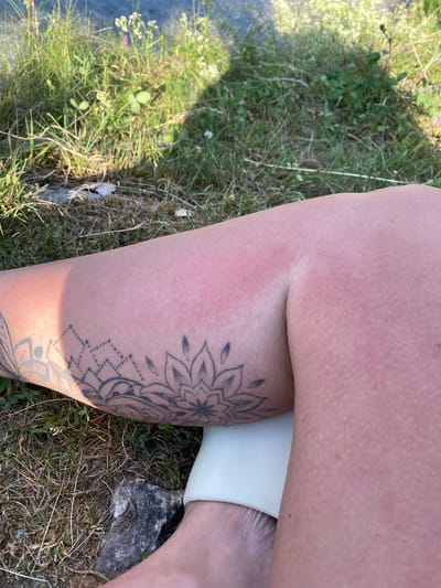 The image shows a close-up view of a person's leg outdoors, with a focus on tanned skin and a tattoo. The skin on the upper part of the leg appears to be sunburned, with a distinct pinkish-red hue, particularly along the side. The lower leg displays a portion of an intricate black tattoo with floral and geometric designs.

The setting is grassy and appears to be a natural environment, possibly a meadow, with dry grass and some green patches. Small white flowers are visible in the background. Sunlight casts a shadow on the leg, suggesting it is daytime. The overall impression is one of being outdoors on a sunny day. There is no visible text in the image. The image seems to capture a moment of relaxation or enjoyment of the outdoors, with the sunburn and tattoo being the primary visual points of interest.