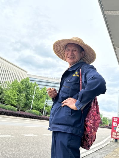 A smiling elderly Asian man wearing a straw hat and a blue work uniform stands on a sidewalk in what appears to be an urban area of China. He has a floral-patterned bag slung over his shoulder. The uniform has text on the front, possibly in Chinese characters. He is holding a lit cigarette in his right hand. Behind him, there is a modern building with a grid-like facade and landscaping with green trees and purple bushes. A street and a sign with Chinese characters on it are also visible. The sky is overcast. The location is inferred to be an unknown city in China.