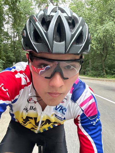 A male individual is depicted in a close-up, first-person perspective, appearing to be engaged in cycling on a paved road. The subject, a young adult, wears a grey and black vented cycling helmet, dark-framed clear cycling glasses with a visible crack on the viewer's left lens and a red reflection on the viewer's right lens. His attire includes a multi-colored cycling jersey, predominantly white, red, and blue, with yellow and black patterns on the lower front. Prominent logos on the jersey include "ia lle", "VKG", "REINIGING & ONDERHOUD", and "MICHELIN". Black cycling shorts are visible below the jersey. A black cord, possibly for earphones, with a small red attachment, is visible around his neck. The subject maintains a neutral expression, looking directly at the camera. The background consists of a grey asphalt road with a white lane line visible on the right, bordered by dense green trees and foliage under natural light, indicative of an outdoor, possibly forested or park-like environment. The scene is located on a road within the city of Bilohorodka, Ukraine.