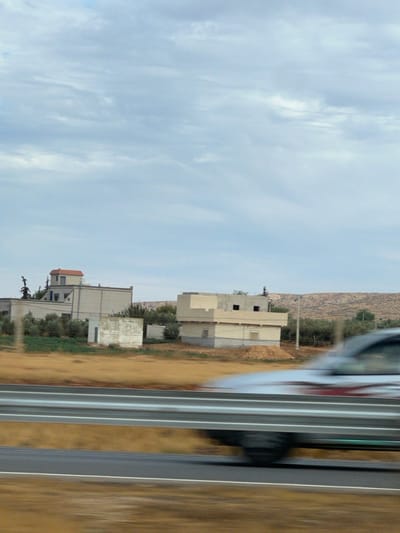 This image, captured in Laqsir, Morocco, presents a roadside view under a partly cloudy sky. In the foreground, a silver-colored vehicle, possibly a truck or SUV, with red and white graphic elements, is in motion from right to left, partially obscured by a two-bar metal guardrail. The road surface, a dark asphalt, is visible beneath the guardrail, transitioning to a strip of dry, yellowish-brown earth.

The midground reveals an arid landscape featuring multiple structures and vegetation. To the left, a light-colored building with a distinct reddish-orange tiled roof structure is visible, accompanied by a smaller, white, rectangular auxiliary building. To the right, another building, appearing unfinished or under construction, displays light beige concrete walls with rectangular openings and is fronted by a mound of reddish-brown earth. Scattered throughout the midground are numerous small trees and green vegetation, interspersed within the dry, light brown soil.

The background consists of a range of barren, rocky hills under a pale blue sky with scattered white and light grey clouds. No individuals are present in the scene.