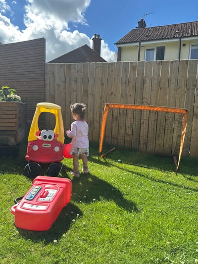 In a sunlit backyard in Belfast City, United Kingdom, a young child stands near a red and yellow toy car and a portable soccer goal. The child, facing away from the camera, is wearing a light pink long-sleeved shirt and patterned shorts. In front of the toy car lies a red toy gas pump. The backyard is enclosed by a wooden fence, with a portion of a residential house visible in the background. The grass is green and appears to be mown, with some small white flowers scattered throughout. The sky is blue with scattered white clouds. The overall scene depicts a playful outdoor environment.