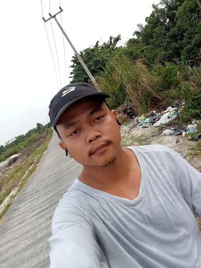 A man with a light complexion, wearing a black baseball cap and a light gray t-shirt, takes a selfie. He has a thin mustache and is wearing black wireless earbuds. He is standing on a paved road with a slight incline, possibly a dike or embankment, with a vegetated area and a pile of garbage to his right. In the background, a telephone pole with wires is visible, along with a dense green forest and a hazy sky. The location is identified as Teluk Kenidai, Indonesia.