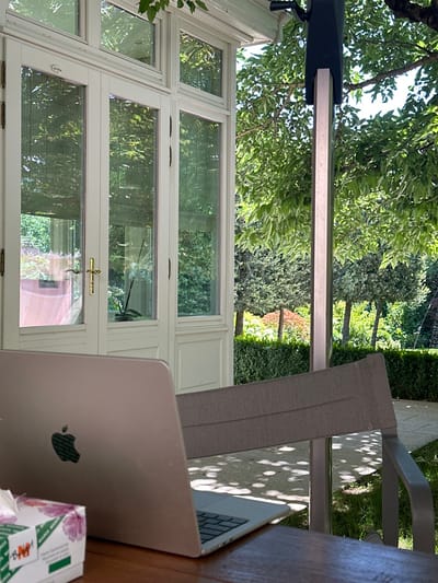 The image depicts an outdoor scene in Vandœuvres, Switzerland, focusing on a personal workspace set up on a patio or terrace adjacent to a building with extensive glasswork. In the foreground, a light brown wooden table holds a light-colored laptop, presented with its lid or back facing the viewer, featuring the visible Apple logo. To the left of the laptop, a white tissue box with a green and pink floral pattern and "BAHH" branding is positioned. Directly behind the table, the back of a grey fabric outdoor chair with a metal frame is partially visible. To the left, a section of a white, multi-paned glass structure, resembling French doors or a large window, forms part of an adjacent building. Golden-colored door handles are present on one of these panels, and through the glass, an orchid plant in a pot is discernible. To the right of the glass structure, a light-colored vertical support beam, possibly metallic, stands, topped with a dark, rectangular mechanism, suggesting an awning or pergola system. The background reveals a lush, sunlit garden with various green trees and sculpted hedges, casting dappled shadows on the ground. A light-colored paved pathway is visible on the far right, leading into the garden area. No individuals are present in the scene.