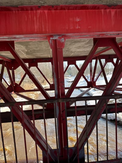 The image displays the underside and structural components of a red metal truss bridge in Oleshkovo, Russia, viewed from below and through a section of its own railing. The primary visible elements are weathered, faded red metal beams and diagonal support structures forming a complex lattice. Paint is peeling in multiple areas, revealing underlying material and showing streaks of discoloration. Above the truss, the concrete underside of the bridge deck is visible, also exhibiting stains and marks.
Through the gaps in the bridge's metal framework and an immediate foreground railing, a river flows, characterized by turbid, brownish-yellow water with visible ripples and current. The river banks on both sides are partially covered with white snow and ice, intermingled with patches of bare earth and dormant vegetation. In the background, numerous bare deciduous trees line the banks under what appears to be a clear sky, indicating a late winter or early spring season. No individuals or active subjects are present in the scene.