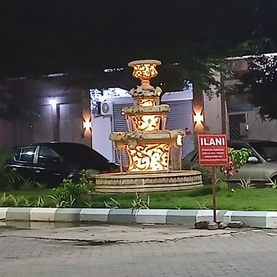The scene depicts an outdoor urban environment at night in Tanga, Tanzania, illuminated by artificial light sources. A large, multi-tiered stone or concrete fountain with intricate cut-out patterns forms the central subject, glowing brightly from internal orange illumination and actively flowing water. The fountain is situated on a circular grassy median containing green foliage. In the background, two vehicles are parked: a dark-colored hatchback or sedan to the left, and a light-colored sedan to the right, both partially obscured by vegetation. Behind the vehicles and fountain, sections of buildings are visible, featuring textured walls, a wall-mounted air conditioning unit, and sconce lighting. A prominent red rectangular sign with white Swahili text, including the word "ILANI," stands on a brown post in the foreground-right. The immediate foreground consists of a cracked, paved surface leading to a curb painted in alternating light and dark grey sections. No individuals or specific actions are depicted.