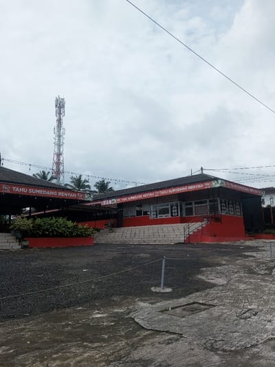 A low-angle shot of a red building with "TAHU SUMEDANG RENYAH" signs around its eaves. The building is tiered, with stairs leading up to its entrance. A telecommunications tower rises in the background, beneath a cloudy sky. A gravel parking area fills the foreground, with patches of concrete.