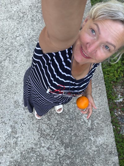 This is a selfie taken outdoors, likely during the daytime with natural lighting. The subject is a woman with blonde hair, wearing a black and white striped dress and white sandals. She is holding an orange in her right hand. She has a bright smile and appears happy. The background consists of a concrete surface, with some greenery and gravel visible at the bottom edge of the frame. The image is a close-up shot focusing on the woman, her outfit, and the orange. There are no visible text elements in the image.