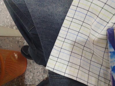 The image shows a close-up view of a person's legs, wearing dark wash denim jeans and a white checkered shirt with thin blue, green, and yellow lines. A portion of an orange, perforated basket is visible on the left side of the frame, and a black shoe is partially visible near the jeans. The ground appears to be a textured surface, possibly concrete or asphalt. The context of Eliowhani, Nigeria, is not discernible from the visual information provided in the image.