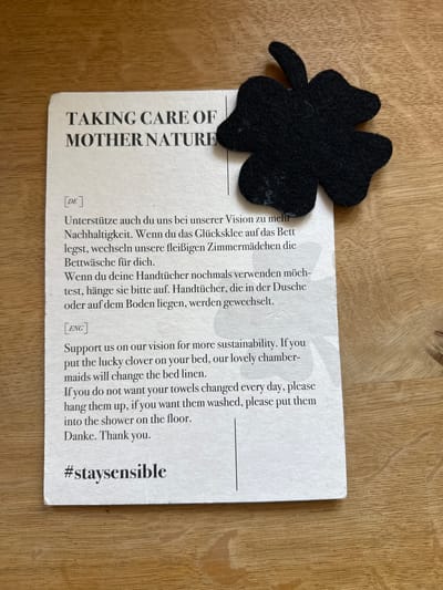 The image shows a white card with black text and a black felt four-leaf clover attached to it. The card has the title "TAKING CARE OF MOTHER NATURE" at the top, followed by text in German and English. Below the English text is the hashtag "#staysensible". The card is placed on a light brown wooden surface. The lighting suggests it is daytime. The scene is an indoor setting, likely a hotel room, given the message about laundry services. There are no people or animals present in the image. The location of St. Märgen, Germany is not indicated in the image.