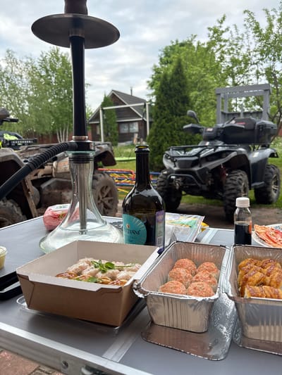 The image shows an outdoor table laden with food containers, a bottle of wine, and a hookah. In the background are two ATVs, one black and one camouflaged, with a small wooden house visible behind the ATVs. The location is Ogryzkovo, Russia.