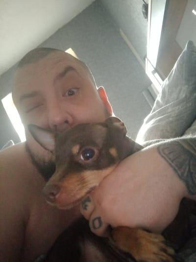 A man with a shaved head and a partial beard is holding a small dog in his arms. The man is winking at the camera, and the dog is looking directly at the viewer with a somewhat concerned expression. The man's arm has a tattoo on it, and there appear to be tattoos on his fingers. The background shows a pillow and a window, suggesting the location is indoors, possibly a bedroom. There is no information in the image to confirm that this image was taken in Chulman, Russia.