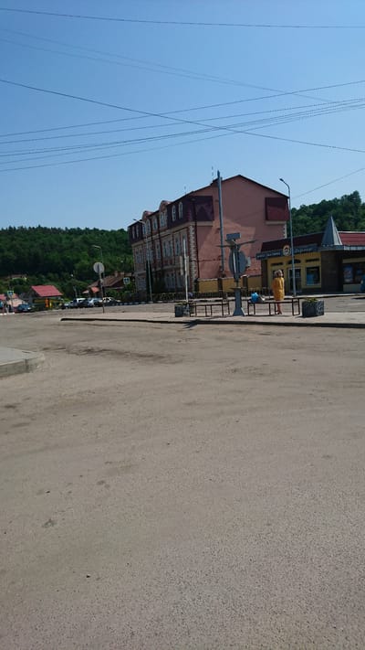 A bright, sunny day with a clear blue sky over a rural town square in Buchach, Ukraine. In the background, a large, multi-story building with a pink facade and a dark red roof stands prominently. Adjacent to it, a smaller building with a yellow facade features signage that appears to read "Фермер" and "МЯСО ЕЖА СВЕЖА". A woman in a yellow dress stands near a bench in front of this building, facing away from the camera. Several cars are parked in the vicinity of the buildings, and a few people are visible in the distance, near a red-roofed structure on the left. The foreground is dominated by a paved road with a slight curve, indicating a street intersection or roundabout. A few streetlights and traffic signs are present in the mid-ground. Green, tree-covered hills form the backdrop to the town.