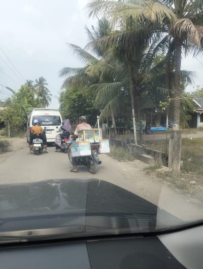 The image depicts a street scene in Teupinreudeup, Indonesia, under an overcast sky, suggesting a warm and humid day. In the foreground, the reflection of palm trees and the sky is visible on the windshield of a vehicle.
The central focus of the image is a traffic jam on a dirt road. A white van is stopped, with text on its rear that appears to be a logo with an airplane and the words "Sili Ayah". Below that, there is a license plate reading "B 0005 FTD".
Several motorcycles are also present in the traffic. One motorcycle in front of the van carries a man wearing an orange shirt and a white cap. Behind the van, two motorcycles are stopped. The first motorcycle has two people on it, a woman in a purple hijab and a man. The second motorcycle, closer to the foreground, is operated by a man wearing a striped shirt and a cap, and it is pulling a cart filled with what appears to be ice cream or similar frozen goods. The cart has text on its side, including the word "JACI" which is partially visible.
The setting is rural, characterized by dense vegetation, including numerous palm trees, and simple buildings with thatched or tiled roofs in the background. A concrete fence runs alongside the road. The overall atmosphere suggests a typical day in a tropical, possibly coastal, Indonesian village. The activity is simply that of vehicles waiting in traffic.
