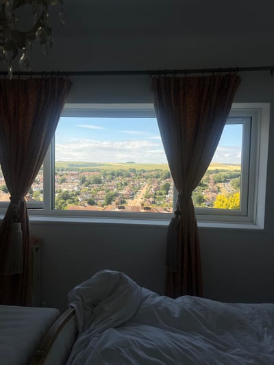 The image shows a view from a window, looking out over a residential area in Brighton, United Kingdom. The window is divided into two panes, and both are open to reveal the scene outside. The top pane looks out onto a green field with some yellow flowers, and the bottom pane shows a town with houses and trees. There are curtains on either side of the window, made of a patterned fabric in shades of brown and gold. The curtains are tied back with tassels.
On the right side of the image, a bed with white bedding is visible. The bed appears to be in a bedroom, and the overall impression is one of a cozy, lived-in space. The lighting suggests it is daytime, likely a sunny day given the bright sky and clear visibility of the scene outside. The time of day is likely late morning or early afternoon. The weather appears to be good, with a clear blue sky and some clouds.
The houses in the town appear to be typical of British architecture, with pitched roofs and brick or tile exteriors. The town is spread out on a gentle slope, and in the distance, a flat, green landscape can be seen. The presence of curtains and a bed suggests this is a domestic setting, likely a home or a hotel room. No people are visible in the image. There is no visible text in the image.