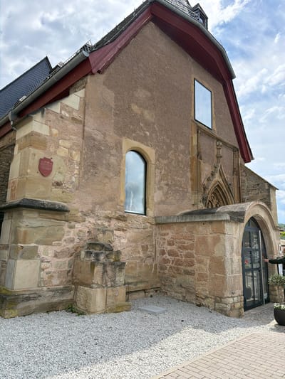 The photograph depicts the exterior of the Synagogue Bad Sobernheim, located in Bad Sobernheim, Germany. The structure, viewed from a low angle, features a blend of architectural styles, with a mix of stone and stucco facades. It incorporates both historical and modern elements. A large, rounded window is integrated into a stone section of the building. Two additional windows, one rectangular and the other arched, are visible on the building's side. A plaque is mounted on the building's facade. The surrounding ground is covered in gravel, and a paved sidewalk borders the building. The sky above is partly cloudy.