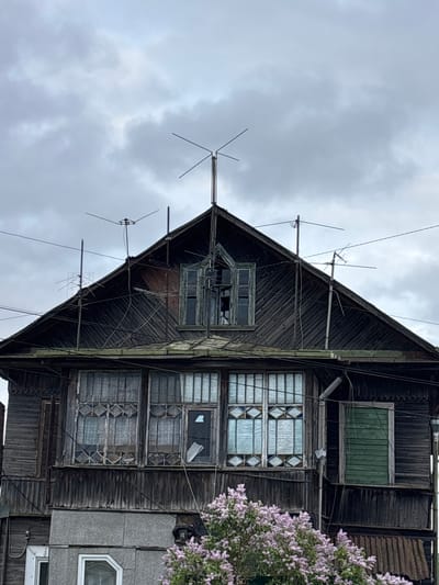 An upward-facing, low-angle shot depicts the gable end of a dilapidated wooden house against a cloudy sky. Several antennas are mounted on the roof and along the top edge of the gable. The dark, weathered wood shows signs of age and disrepair. The windows are a mix of styles; some have a decorative diamond pattern, while others are boarded up or have broken panes. To the right of the house, a flowering bush with purple blossoms is visible. A section of a more modern building with a gray exterior adjoins the wooden structure. There is no indication of people or specific actions in progress. The scene appears to be in a state of decay and neglect, possibly in a rural or less affluent area. Ulyanovka, Russia is not evident in the image.