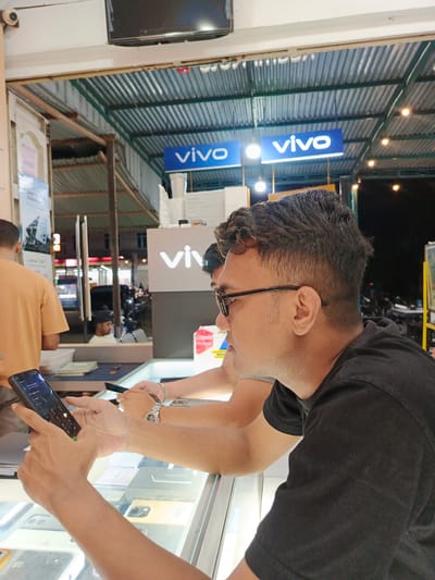 The image depicts an indoor scene at a mobile phone store, possibly located in Namplohbaro, Indonesia, indicated by the "vivo" branding prominently displayed. Two men are the primary subjects. In the foreground, a man wearing a black t-shirt and glasses is holding a black smartphone, seemingly interacting with its screen. His right arm rests on a glass display counter, which exhibits various mobile phones and boxes. To his left, another man, wearing a tan t-shirt, is partially visible, also appearing to be engaged with the display. The background reveals more of the store's interior, including a corrugated metal ceiling and additional "vivo" signage. The overall lighting suggests it is either evening or a dimly lit interior space.