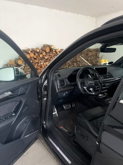 The image shows the interior of a black car, likely an Audi SQ5, with a view of the steering wheel, dashboard, and front seats. Wood logs are stacked outside the driver's side door. The vehicle appears to be parked indoors, possibly in a garage with white walls. The lighting suggests it may be daytime.