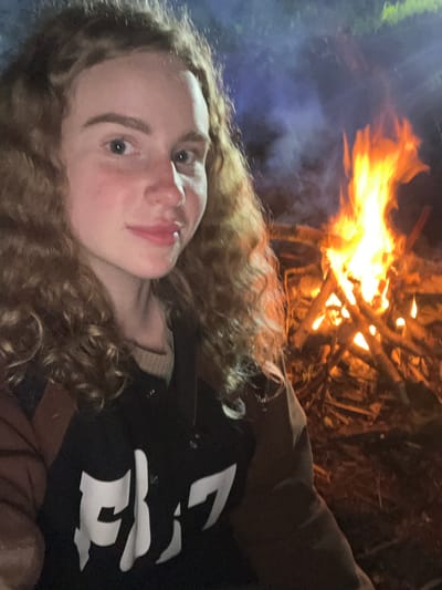A young woman with curly hair is facing the camera in a selfie. She is wearing a black jacket with large white lettering. Behind her is a campfire, with flames and smoke visible. The location is unknown. Krasnyi Jar, Russia is not discernible.