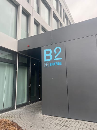 The image presents a modern building entrance in Meyrin, Switzerland, viewed on an overcast day. A prominent dark gray panel extends from the building, serving as a canopy over the entrance. On this panel, light blue signage is clearly visible, featuring the large characters "B2" and the vertically oriented word "ENTREE" (French for Entrance), accompanied by an upward arrow symbol in a square box, indicating the entry point.

The building exhibits contemporary architecture, characterized by a light-colored facade composed of a grid of horizontal and diagonal framing elements enclosing multiple large, reflective windows. These windows mirror the muted, cloudy sky, creating a soft, diffused lighting effect. Below the dark gray canopy, glass entrance doors are partially visible, offering a glimpse into the building's interior. A small, red circular object, possibly a sensor or indicator light, is affixed to the building's wall near the entrance.

The ground surrounding the entrance is paved with small, light-toned cobblestones. In the bottom right corner, some dry, brown vegetation or bushes are visible. The scene is devoid of people, emphasizing the architectural features and the functional nature of the entrance. The consistent soft light suggests it is daytime without direct sunlight.