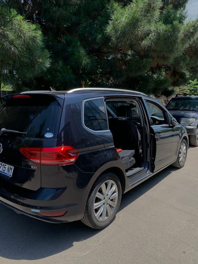 A dark blue Volkswagen Touran is parked on an asphalt surface, with its rear passenger door and driver's side door open. The vehicle is positioned at an angle, facing slightly away from the viewer, with the rear of the car on the left side of the frame and the front extending towards the right. The interior of the car is visible, showing folded down seats and a red object on one of the seats. A dark-colored SUV is parked behind the Volkswagen. The background consists of lush green pine trees and a bright, overcast sky. A license plate on the Volkswagen reads "715 NM". A sticker on the rear window of the Volkswagen displays Japanese text and stars. Another sticker on the rear bumper reads "AOI Euro". The scene appears to be in an outdoor parking area, possibly in Rustavi, Georgia, given the presence of Japanese vehicle stickers, which are common in areas with a significant Japanese car import market.