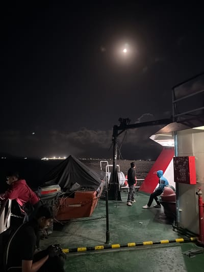The image depicts a nighttime scene aboard a ferry or ship, likely near the coast of Ketapang, Indonesia, given the distant city lights and cloudy sky. Four individuals are present on the deck. In the foreground left, a person in a red hoodie leans over a railing. To their right and slightly further back, another person in a black shirt is seated, looking down at something in their hands, possibly a camera. In the mid-ground, two more individuals are standing. One, wearing a dark hooded jacket, looks out towards the water. The other, in a blue hooded jacket, is seated on a rounded object. The deck is green and marked with yellow and black striped safety barriers. A covered life raft or similar vessel is visible, with an outboard motor attached. A crane-like structure extends over the deck. The moon is bright in the dark, cloudy sky, and distant lights from a city or port are visible on the horizon over the water.