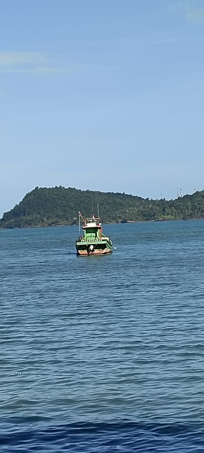 A green and brown fishing boat is in the middle of a calm blue sea. In the background, a lush green island with many trees rises from the water, its slopes covered in dense vegetation. The sky above is a clear, pale blue with a few wispy white clouds. The scene appears to be captured on a bright, sunny day. The location is Lhoktimon, Indonesia.