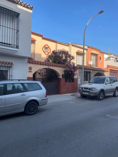 The image shows a street scene in La Línea de la Concepción, Spain. Two silver cars are parked on the side of a paved street. The car in the foreground is a hatchback, and the car in the background is a larger SUV. The buildings lining the street are residential, with terracotta tile roofs and pastel-colored facades. One building features decorative tile work around a window and an arched entryway with a dark metal gate. A flowering tree with pink blossoms is visible in front of one of the buildings. A street lamp extends over the sidewalk. The sky is clear and blue, suggesting it is daytime. The lighting indicates it is likely late afternoon or early evening. There is some visible text on the license plate of the SUV, which reads "7209 BLZ". There is also a circular orange sign on one of the buildings with black text, which is not legible.