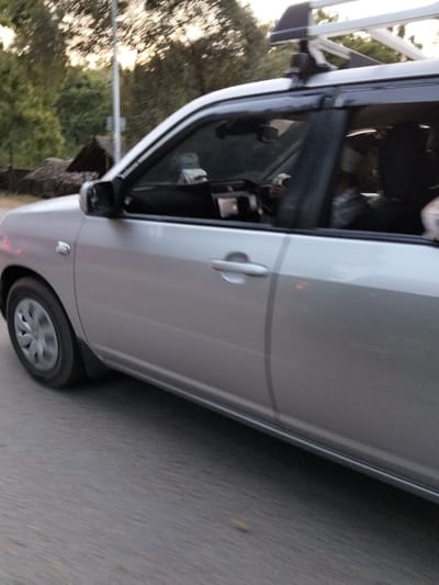 The image displays a silver-colored vehicle, likely a station wagon or small SUV, in motion on a paved road within the Gombato Bongwe ward, Kenya. The vehicle is seen from its front-left side, showing the driver's side door, front fender, and partially obscured front wheel. A silver roof rack with black supports is installed on the vehicle's roof, and dark rain deflectors are present above the side windows. The road surface is dark grey and exhibits horizontal motion blur, indicating vehicular movement.

Inside the vehicle, a driver is visible, positioned on the right side of the vehicle (consistent with left-hand drive traffic). The driver's hands are on the steering wheel, and they appear to be wearing a light-colored top. A front passenger is partially visible as a dark silhouette to the driver's left. A rear passenger's dark silhouette is also partially visible behind the driver. The interior shows reflections on the windshield and some dashboard elements.

The background features green trees and foliage, with a utility pole rising vertically behind the vehicle's front door. To the far left, a structure with a dark brown, possibly thatched or corrugated iron, roof is partially visible amidst the foliage. The lighting suggests daylight conditions.