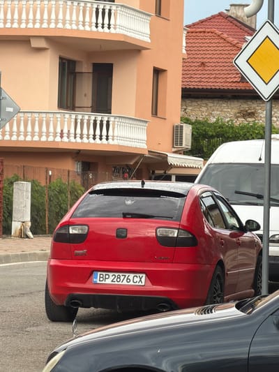 The image shows a street scene in Hadzhidimovo, Bulgaria. The primary subjects are several parked cars, with a bright red hatchback in the foreground. The hatchback's rear end is prominent, displaying a license plate that reads "BP 2876 CX". To the right of the red car is a dark-colored vehicle, likely a sedan or SUV, reflecting the surroundings. In the background, a white van is partially visible.
The setting appears to be a residential or commercial area. A building with peach-colored walls and white ornate balconies with balustrades stands to the left of the cars. The building has several windows, some of which are dark. Above the red car, a roof with red tiles is visible. A street sign with a yellow diamond shape on a white background is also present in the upper left corner.
The image is taken from a slightly elevated perspective, looking down towards the street and cars. The lighting suggests it might be daytime, possibly overcast or with diffused light. There are no people visible in the frame. The overall mood is one of stillness, characteristic of a parked scene on a street. The focus is on the vehicles and the immediate architectural surroundings.