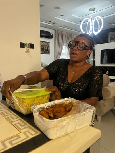 An older woman with glasses is shown standing near a table inside a house in Mopo-Akinlade, Nigeria. She wears a black top and jewelry. Two covered containers, one with yellow rice and the other containing chicken, rest on the table beside her. The table has a black and beige pattern around the edges. In the background, there is a modern chandelier, a wall with a picture frame and a ventilation unit, and a partially visible couch.