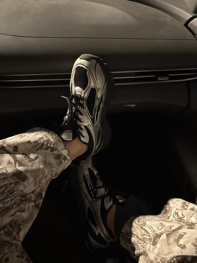Two sneakers are positioned within a dark vehicle interior. The left shoe rests on the dashboard, while the right shoe is on the floor. Both are white and black with laces. The person wearing the shoes is also wearing patterned beige camouflage pants. The lighting is dim.
